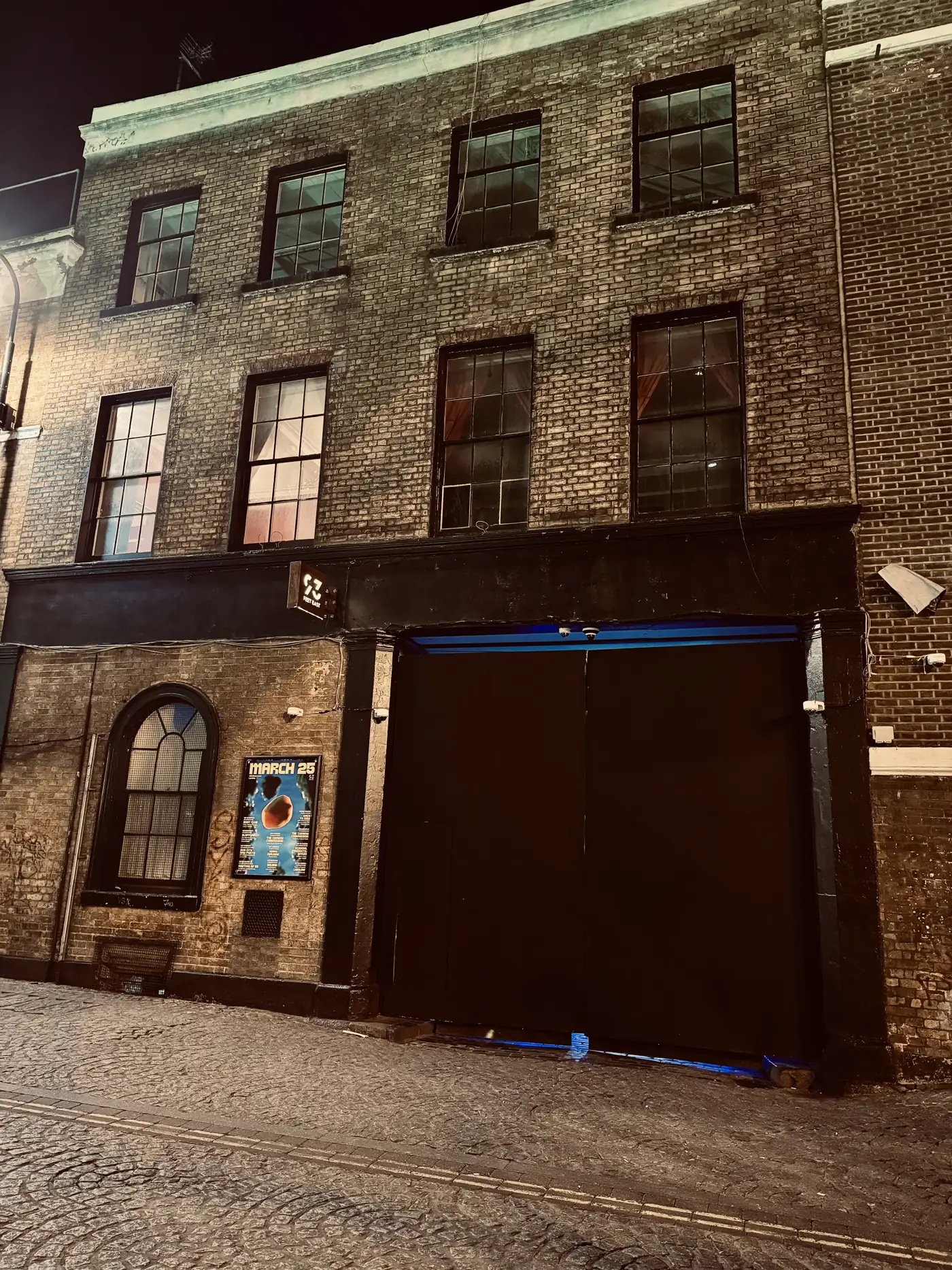 The black door of a studio on Brick Lane at night, blue light leaking from beneath it.